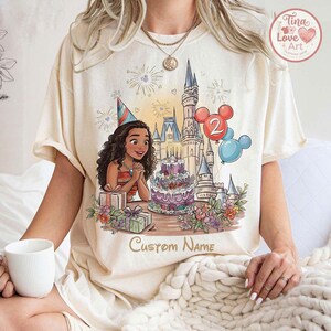 May include: Cream-coloured t-shirt with a cartoon illustration of a birthday scene. The design includes a character wearing a party hat, a castle, a cake, balloons, and the text "Custom Name". The shirt has short sleeves.