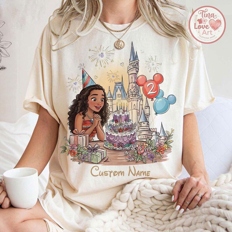 May include: Cream-coloured t-shirt with a cartoon illustration of a birthday scene. The design includes a character wearing a party hat, a castle, a cake, balloons, and the text "Custom Name". The shirt has short sleeves.