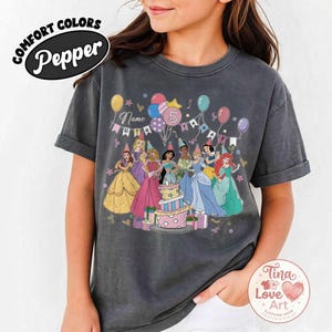 May include: Dark gray t-shirt featuring a colorful graphic of Disney princesses around a birthday cake. The design includes balloons, the number 5, and the text "Name Birthday". The shirt has rolled-up sleeves and the words "Comfort Colors Pepper" are printed on the top left.