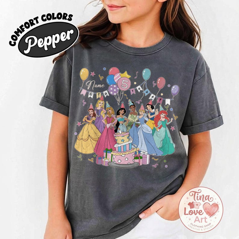 May include: Dark gray t-shirt featuring a colorful graphic of Disney princesses around a birthday cake. The design includes balloons, the number 5, and the text "Name Birthday". The shirt has rolled-up sleeves and the words "Comfort Colors Pepper" are printed on the top left.