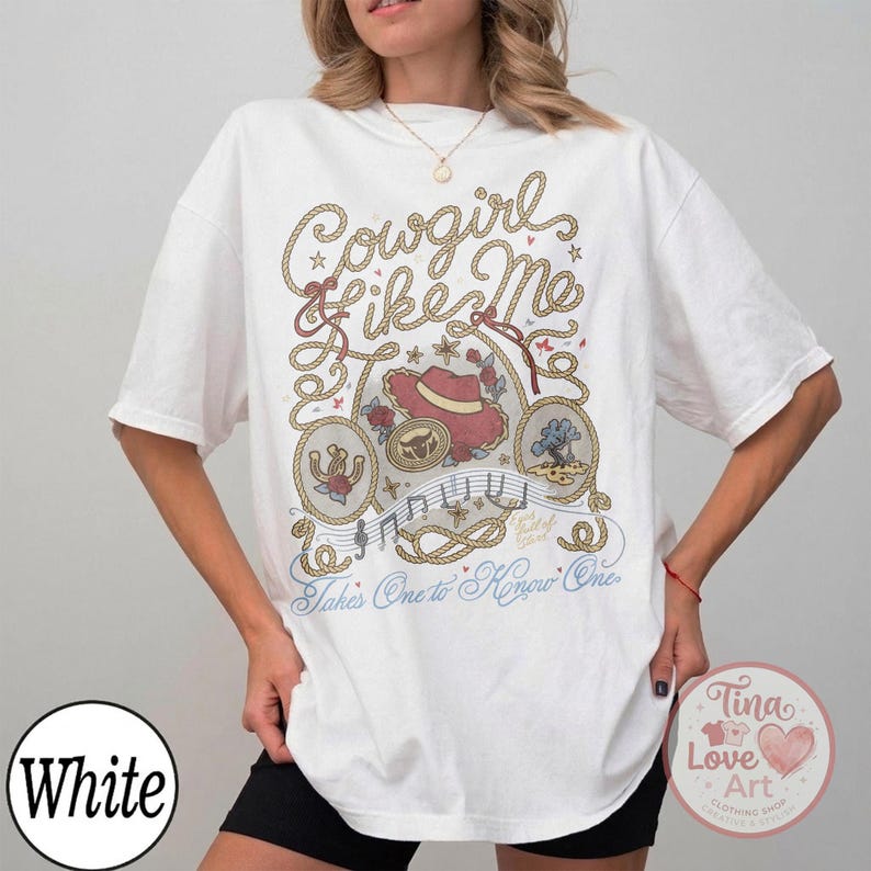 Pu&ograve; includere: Una t-shirt bianca a maniche corte con una stampa grafica bordata da una corda, un cappello da cowboy rosso, un ferro di cavallo e un paesaggio desertico. Il testo recita Cowgirl Like Me e Takes One to Know One in caratteri marroni, rossi e blu.