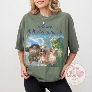 May include: An olive green t-shirt featuring the Disney movie "Moana." The graphic includes the movie title in blue and the characters Moana, Maui, and others. The shirt is a relaxed fit.