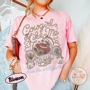 Pu&ograve; includere: Una t-shirt a maniche corte rosa chiaro con una stampa grafica raffigurante un design a corda da lazo, un cappello da cowboy, un ferro di cavallo e note musicali. Il testo recita Cowgirl Like Me e Takes One to Know One in corsivo.