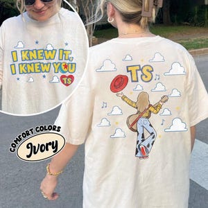 K&ouml;nnte beinhalten: Ein elfenbeinfarbenes T-Shirt mit Grafikdruck. Die Vorderseite zeigt den Text I KNEW IT, I KNEW YOU TS in blauen Buchstaben, ein rotes Herz und Sterne. Die R&uuml;ckseite zeigt eine Person mit Gitarre, rotem Hut und Hose mit Kuhfleckenmuster.