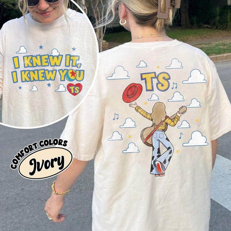 K&ouml;nnte beinhalten: Ein elfenbeinfarbenes T-Shirt mit Grafikdruck. Die Vorderseite zeigt den Text I KNEW IT, I KNEW YOU TS in blauen Buchstaben, ein rotes Herz und Sterne. Die R&uuml;ckseite zeigt eine Person mit Gitarre, rotem Hut und Hose mit Kuhfleckenmuster.