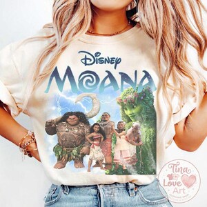 May include: Cream-colored t-shirt featuring the Disney movie "Moana" graphic. The design includes the movie title, characters, and a lightning bolt background. The shirt has short sleeves and a crew neck.