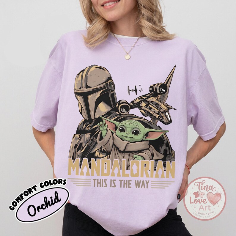 Pu&ograve; includere: T-shirt color orchidea con una grafica di The Mandalorian, Grogu e un'astronave. I testi "MANDALORIAN" e "THIS IS THE WAY" sono stampati in oro. La maglietta ha un'etichetta Comfort Colors.