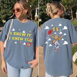 K&ouml;nnte beinhalten: Ein blaues T-Shirt mit Grafikdruck. Die Vorderseite zeigt den Text I KNEW IT, I KNEW YOU mit einem Ball-Symbol und einem roten Herz mit den Buchstaben TS. Die R&uuml;ckseite zeigt eine Person mit Gitarre, rotem Hut und den Buchstaben TS zwischen Wolken.