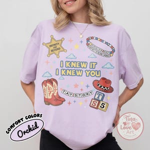 May include: A light purple t-shirt featuring a graphic print with a yellow sheriff star labeled Infinity and Beyond, a beaded bracelet, cowboy boots, a red hat, and blocks with the text TAY STORY and I KNEW IT I KNEW YOU with colorful stars.