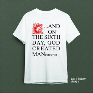 Könnte beinhalten: Weißes T-Shirt mit dem Text "...AND ON THE SIXTH DAY, GOD CREATED MANCHESTER" in schwarzer Schrift. Das Design enthält eine rote dekorative Grafik. Das Shirt hat einen Rundhalsausschnitt.