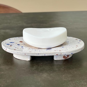 Puede incluir: Una pastilla de jabón blanca descansa sobre un jabonera de cerámica moteada. La jabonera tiene forma ovalada con base blanca y motas azules y marrones. La jabonera tiene una plataforma elevada.