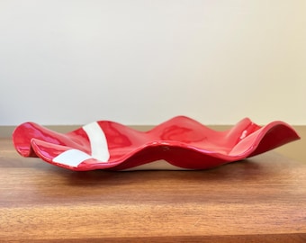 Könnte beinhalten: Eine glänzende rote Keramikservierschale mit gewelltem, gerüschtem Rand. Ein weißer Streifen akzentuiert die Oberfläche der Schale. Die Schale wird auf einer Holzoberfläche präsentiert.