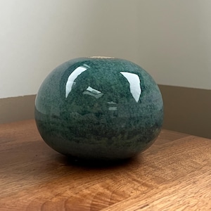 Op de afbeelding: Een kleine, ronde, teal-kleurige keramische vaas met een glanzende afwerking. De vaas heeft een enkele opening aan de bovenkant en staat op een houten oppervlak. De teal kleur heeft een gespikkelde uitstraling.
