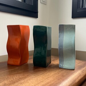 Peut inclure: Trois vases en céramique. Le premier est orange, le second vert foncé et le troisième bleu clair avec des rayures grises. Chaque vase a une forme rectangulaire avec un design légèrement ondulé.