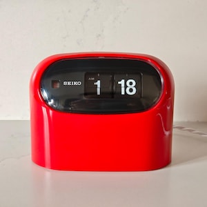 May include: A red Seiko flip clock displaying the time as 1:18 AM. The clock has a glossy finish and a black display window. The word "SEIKO" is printed on the top left of the clock face.