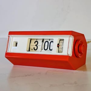 Puede incluir: Un reloj de solapa rectangular de color rojo brillante con una cara blanca y números negros que muestran las 3:00 PM. El reloj cuenta con una cubierta de plástico transparente sobre la pantalla, un dial rojo circular y un cable beige.