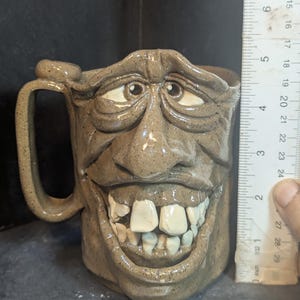 May include: A handmade ceramic mug with a sculpted face design. The mug is approximately 4 inches tall, featuring a wide, toothy grin, and detailed facial features. The mug has a handle and is finished in a speckled, earthy brown glaze.