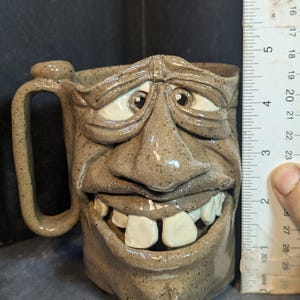 May include: A handmade ceramic mug with a face design. The mug is approximately 4 inches tall. It features a detailed face with a wide grin, large teeth, and expressive eyes. The mug has a speckled, earthy brown color and a handle on the side.