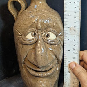 Puede incluir: Una jarra de cerámica marrón con un diseño de cara sonriente. La jarra tiene un asa y un pico. La cara tiene ojos pintados de blanco y una nariz grande. La jarra mide aproximadamente 20 cm de alto.