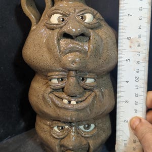 Könnte beinhalten: Ein brauner Keramikkanne mit Henkel, die drei gestapelte Gesichter mit übertriebenen Ausdrücken zeigt. Die Kanne ist etwa 20 cm hoch, wobei jedes Gesicht einzigartige Merkmale und Ausdrücke aufweist. Die Gesichter haben weiße Augen und bestehen aus einem strukturierten, erdigen Material.