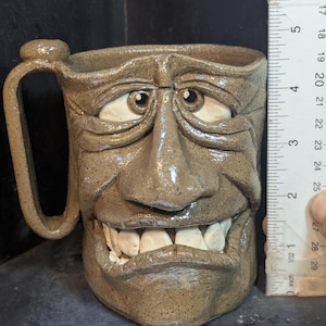 May include: A handmade ceramic mug with a sculpted face design. The mug is approximately 4 inches tall and features a handle. The face has detailed eyes, a nose, and a wide, toothy grin. The mug is a neutral brown color.