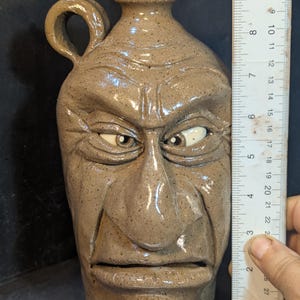 May include: A stoneware jug with a face sculpted on the front. The jug is tan with dark speckles and has a handle on the side. The face has furrowed brows, a stern expression, and measures approximately 10 inches in height.