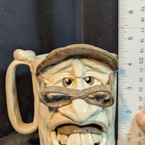 May include: A handmade ceramic mug with a sculpted face design. The mug features a handle and is approximately 5 inches tall. The face has a hat, glasses, and a wide open mouth. The mug is off-white with brown accents.