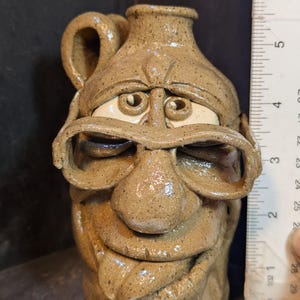 May include: A tan ceramic jug with a face sculpted on the front. The jug has a handle and a spout. The face has large eyes, a nose, and a smiling mouth. The jug is approximately 5 inches tall.
