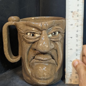 May include: A stoneware mug with a sculpted face design. The mug is approximately 4 inches tall, with a handle and a detailed, expressive face. The color is a speckled brown.