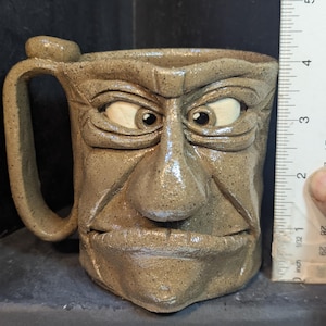 May include: A handmade stoneware mug with a sculpted face design. The mug is approximately 4 inches tall and features a handle. The face has detailed features, including eyes, a nose, and a mouth. The mug is a neutral tan color.