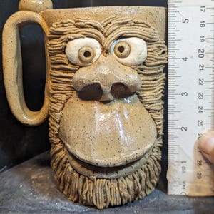 Op de afbeelding: Een handgemaakte keramische mok met een gebeeldhouwd aapachtig gezicht. De mok is bruin met witte accenten voor de ogen en heeft een groot handvat. De mok is ongeveer 10 cm hoog.