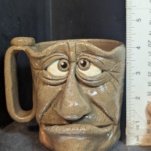 May include: A handmade ceramic mug with a sculpted face design. The mug is approximately 3 inches tall and features a handle. The face has detailed features, including eyes, a nose, and a mouth. The mug is a light brown color.