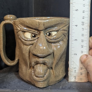 May include: A handmade ceramic mug with a sculpted face design. The mug is approximately 4 inches tall and features a handle and a detailed face with bulging eyes and a protruding tongue. The mug is a speckled brown color.