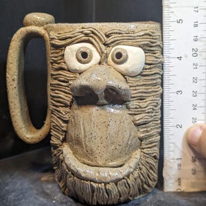 Op de afbeelding: Een handgemaakte keramische mok met een grillig apengezichtontwerp. De mok is ongeveer 10 cm hoog en heeft een getextureerde, gespikkelde afwerking. Het handvat is gebogen en het gezicht heeft grote, expressieve ogen en een prominente neus.