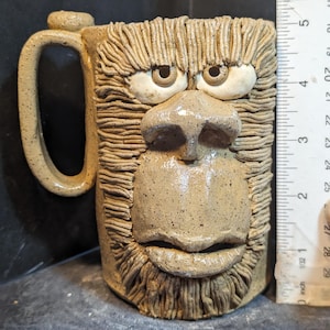 Op de afbeelding: Een handgemaakte keramische mok met een gebeeldhouwd gezicht. De mok is bruin met een textuur, met een grote neus, ogen en een mond. De handgreep is een eenvoudige lus. De mok is ongeveer 10 cm hoog.