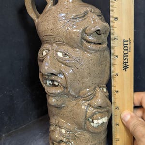 May include: A brown speckled ceramic face jug featuring four stacked sculpted faces with distinct expressions and visible teeth. A wooden Westcott ruler placed vertically next to the jug indicates a height of approximately 25.4 cm for the vessel.