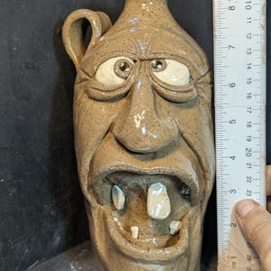 Op de afbeelding: Een bruin keramieken kruik met een gezicht gebeeldhouwd op de voorkant. De kruik heeft een handvat en een tuit. Het gezicht heeft grote ogen, een wijd open mond met tanden en een gefronste wenkbrauw. De kruik is ongeveer 20 cm hoog.