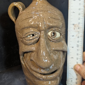 Peut inclure: Une cruche en céramique beige avec un visage sculpté. La cruche a une anse et un col étroit. Le visage a un œil fermé et l'autre ouvert, avec une large bouche souriante. La cruche mesure environ 25 cm de haut.