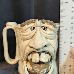 May include: A ceramic mug with a sculpted face design. The mug is off-white with brown accents for the eyebrows, tongue, and teeth. The mug is approximately 5 inches tall.