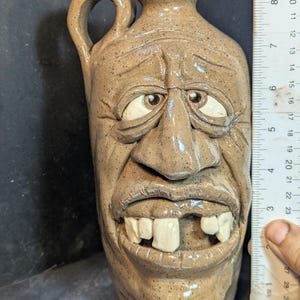 Puede incluir: Una jarra de cerámica marrón con asa, esculpida con una cara. La cara tiene ojos grandes y expresivos, una nariz ancha y una sonrisa con dientes. La jarra mide aproximadamente 20 cm de altura.