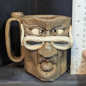 May include: A handmade ceramic mug with a face design, featuring a handle and a small knob. The mug is approximately 4 inches tall. The face has white glasses, eyes, a nose, and a mouth. The mug is a light brown color.