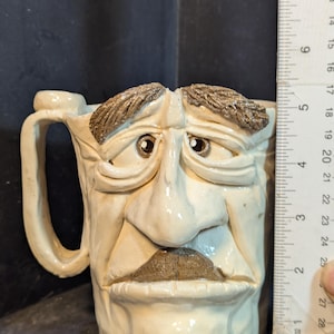 May include: A ceramic mug with a sculpted face design. The mug is off-white with brown accents for the eyebrows and mustache. The mug is approximately 4 inches tall, as measured by the ruler in the image.