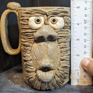 Op de afbeelding: Handgemaakte keramische mok met een gebeeldhouwd gezicht. De mok heeft grote, expressieve ogen, een neus en een mond, allemaal in een getextureerde, aardse tint. De mok is ongeveer 10 cm hoog, met een handvat aan de zijkant.