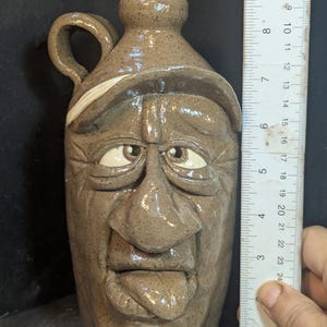 May include: A stoneware jug with a face sculpted on the front. The jug is approximately 10 inches tall. The face has a hat, eyes, nose, and a tongue sticking out. The jug is a light brown color.