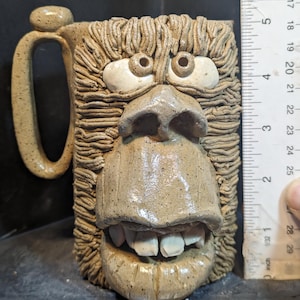Op de afbeelding: Een handgemaakte keramische mok met een gebeeldhouwd gezichtsontwerp. De mok heeft een gedetailleerd, getextureerd oppervlak met een lichtbruine kleur. Het gezicht heeft grote ogen, een prominente neus en een brede, tandige grijns. De mok is ongeveer 10 cm hoog.