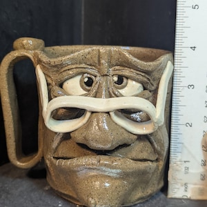 May include: A handmade ceramic mug with a sculpted face design. The mug is tan with white-rimmed glasses and a handle. The mug is approximately 4 inches tall, as measured by the ruler in the image. The mug has a unique, artistic appearance.