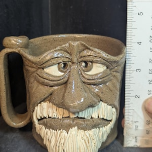 May include: A handmade ceramic mug with a sculpted face design. The mug is a speckled brown color with a handle on the side. The face has white teeth and a white beard. The mug is approximately 4 inches tall.