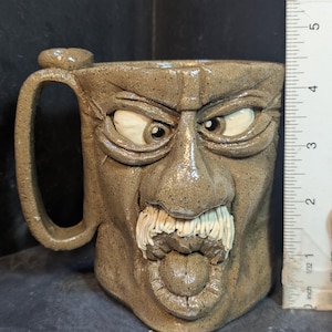 May include: A handmade ceramic mug with a sculpted face design. The mug is a speckled tan color with a handle and a small thumb rest. The face features bulging eyes, a mustache, and an open mouth with teeth and a tongue.