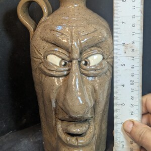 May include: A tan ceramic jug with a handle, featuring a sculpted face with wide eyes and a grinning mouth. The jug is approximately 23 cm tall. The jug has a speckled texture and a rustic appearance.