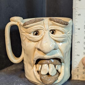 May include: A handmade ceramic mug with a sculpted face design. The mug is off-white with brown accents on the eyebrows, eyes, and tongue. The mug is approximately 4 inches tall.